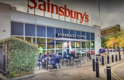 Starbucks: Outdoor seating area with black metal tables and chairs, two large umbrellas, two people sitting and a large glass storefront with Starbucks Coffee sign.