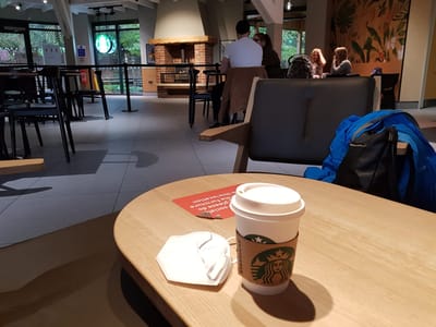Starbucks Lakeside: Round wooden table with a takeaway coffee cup and a mask, black cushioned armchair nearby, and a few people seated at tables in the background.