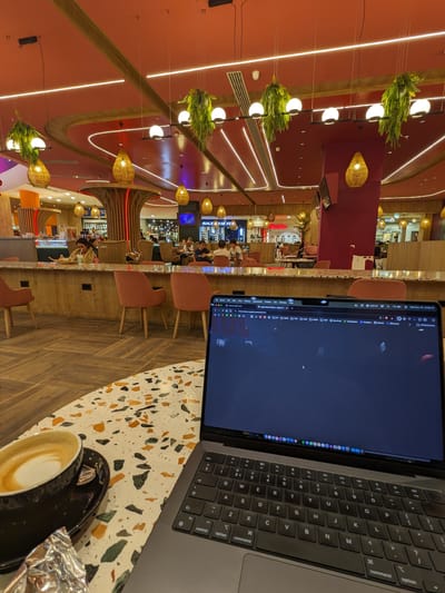 The Point Shopping Mall: Terrazzo table with a laptop and coffee cup, looking out to a long counter with pink chairs and hanging wicker and globe lights above.