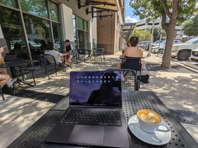Merit Coffee: Metal tables and chairs on a shaded sidewalk patio with a laptop and latte on one table and several people seated at others.