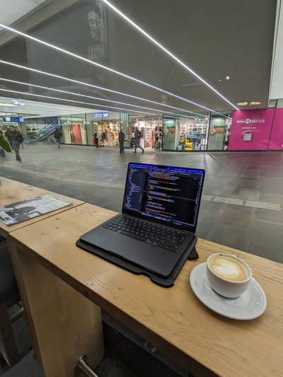 Coffee Fellows Eindhoven Centraal: Long wooden counter with a laptop, magazine, and a cup of coffee overlooking a spacious tiled shopping area with several people walking.