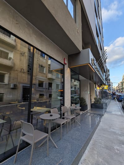 Regional Hotel: Three small round tables with plastic chairs set outside along a building with large reflective windows and a covered entrance.
