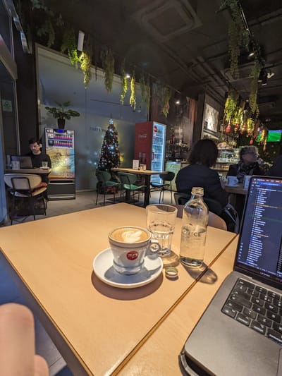 Cafe Pascucci: Light wooden tables with a coffee cup, glass, and bottle, surrounded by metal chairs, a Christmas tree, and a few people working on laptops.