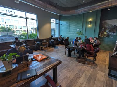 Bennu Coffee: Two large wooden tables with chairs and desk lamps, bench seating along windows with small tables, several people working on laptops.