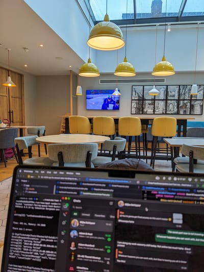 Hampton by Hilton Canterbury: Round wooden tables with grey chairs in front of a high table with six yellow stools under yellow and white hanging lamps and a TV.