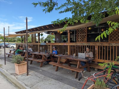 Epoch Coffee: Two wooden picnic tables with benches on a paved area, one person using a laptop, and a covered wooden deck with lattice and hanging lights.