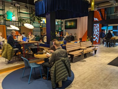 StartHub Cafe: Round wooden table with blue chairs and a jacket on one, adjacent to a long wooden bench with small attached round tables, several people working.