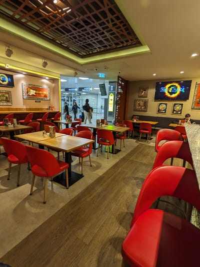 Hard Rock Cafe: Red cushioned chairs around wooden tables and a marble counter with red bar stools, some people walking near an airport entrance in the background.