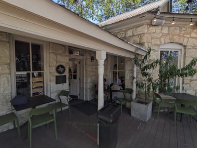 Merit Coffee: Stone building porch with four green chairs at three small dark tables, a trash bin, two potted plants, and people seated inside by a window.