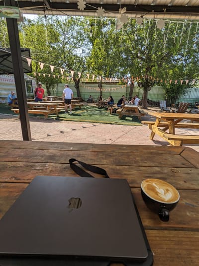Barrett’s Too (Formerly Double Trouble): Wooden picnic tables spread across a patio and artificial grass area with bunting and string lights overhead, a laptop and coffee cup on a table in the foreground.