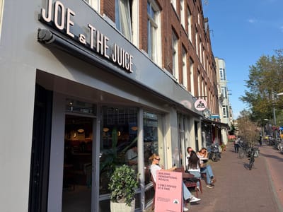Joe & The Juice: Storefront with large JOE & THE JUICE sign, pink sandwich board offering free coffee, and outdoor seating with four people.