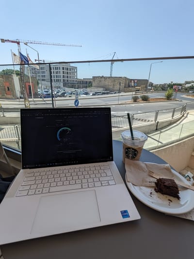 Starbucks: Laptop on a round table by a window with a plastic cup of iced coffee and a plate with a brownie on a napkin.