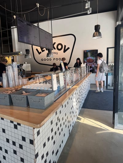 Lebkov & Sons Leiden: Tiled counter with a wood edge holding utensils and cups, a large hanging menu, and three staff behind serving a customer near the entrance.