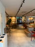 Long narrow seating area with small round tables, wooden chairs, a bench with cushions, and plants along the walls.