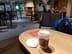 Starbucks Lakeside: Round wooden table with a takeaway coffee cup and a mask, black cushioned armchair nearby, and a few people seated at tables in the background.