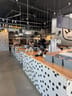 Long counter with a wood top and black-and-white tiled front, three industrial pendant lights above, and a few people seated at tables behind it.