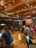 Wooden stools and clear plastic chairs at small tables with laptops, exposed ducts and string lights overhead, brick wall with coat rack and exit door.