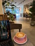 Round table with a laptop and a latte in a pink cup, wooden chairs, large potted plants, and a person working near the back wall with framed art.