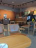 Starbucks: Wooden tables with padded chairs and armrests, a few people standing near the counter, and a small cabinet with notices on the wall.