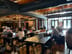 Large wooden tables with chairs and a sofa, several people working on laptops, exposed ductwork and string lights overhead, bright entrance doors.