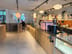 Counter with coffee machine, grinder, and glass display, with a tall wooden table and stools near three mirrors on the wall.