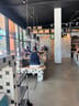 Wooden benches and tables with tiled bases, three hanging lamps above, a few people seated inside a bright cafe with large windows.