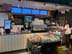 Counter with a coffee machine, cups, display cases of food, refrigerated drinks, digital menus, and a person ordering.